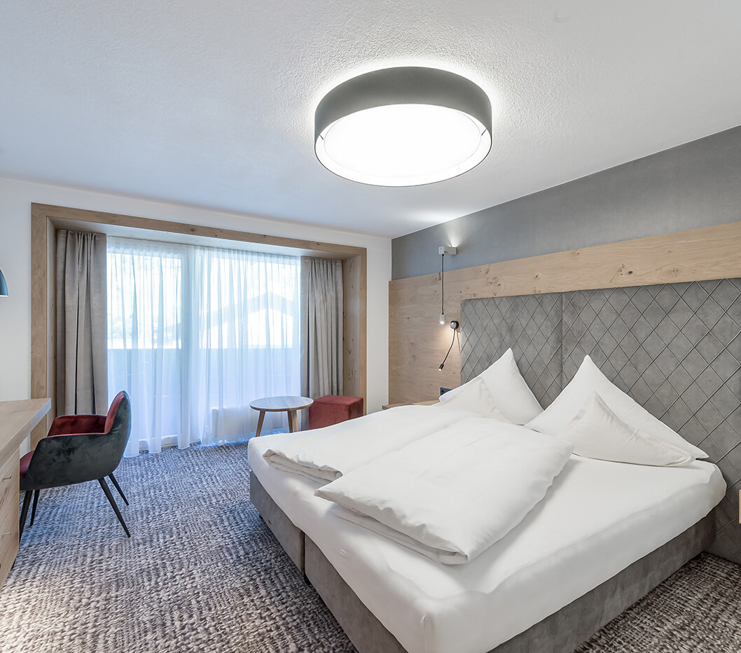 Modern hotel room with a large bed, wooden furniture, a desk with a chair, a small round table and a window with sheer curtains that let in daylight.