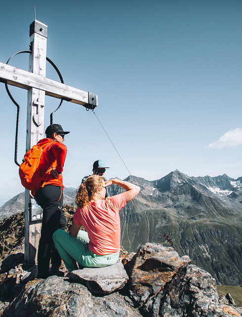 Zwei Wanderer ruhen sich neben einem großen Holzkreuz auf einem Berggipfel aus und genießen den malerischen Ausblick auf die schroffen, weit entfernten Gipfel unter einem strahlend blauen Himmel.